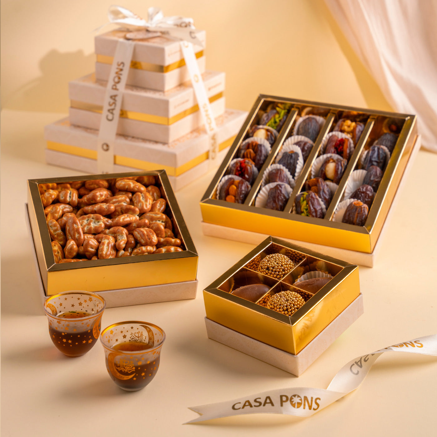 An assortment of luxury food items in beautifully designed white and gold colored boxes, labeled as 'CASA PONS'. The items include dates with nuts, chocolate dates, and other confectionary products.