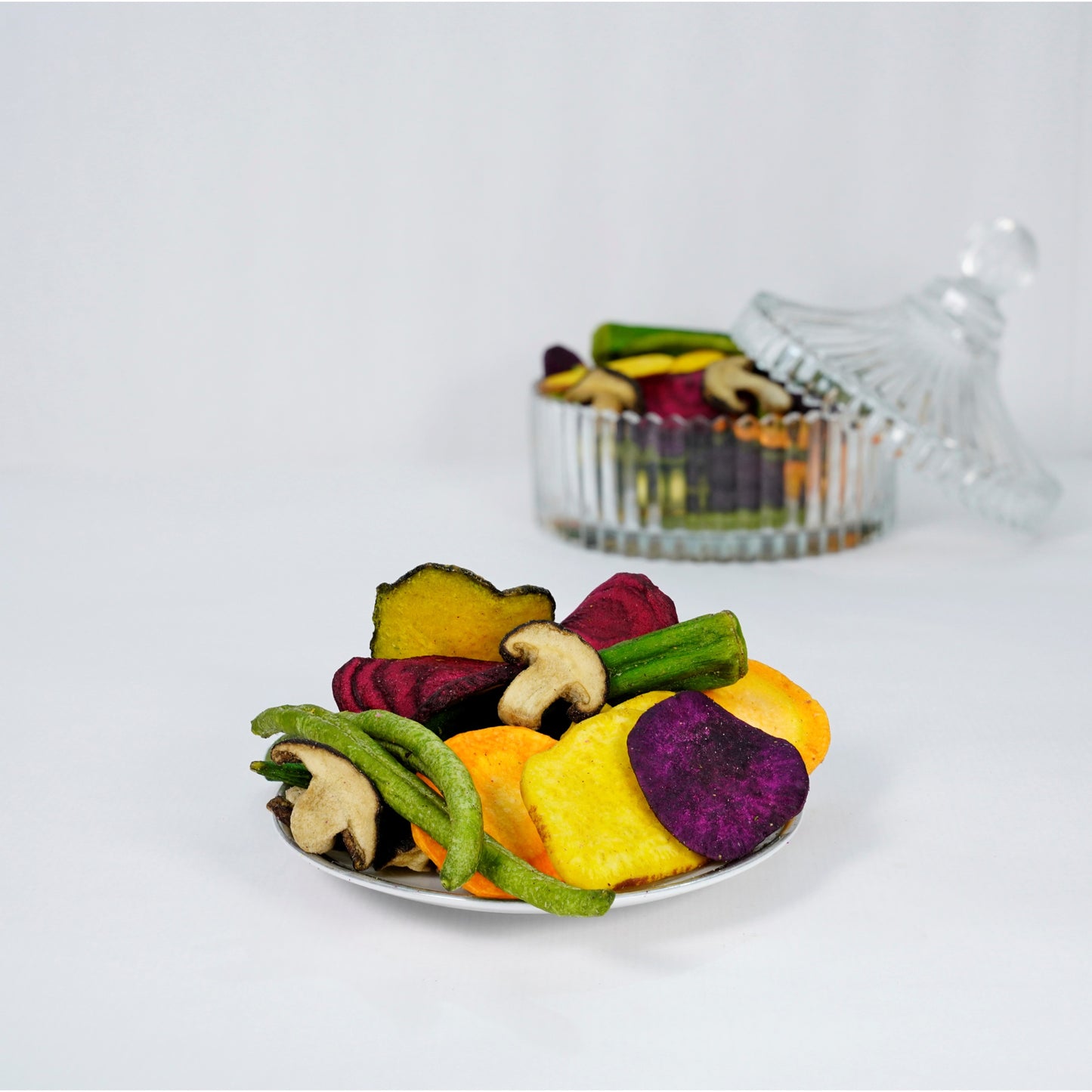 Colorful dried vegetable chips on a white plate with a clear container in the background.