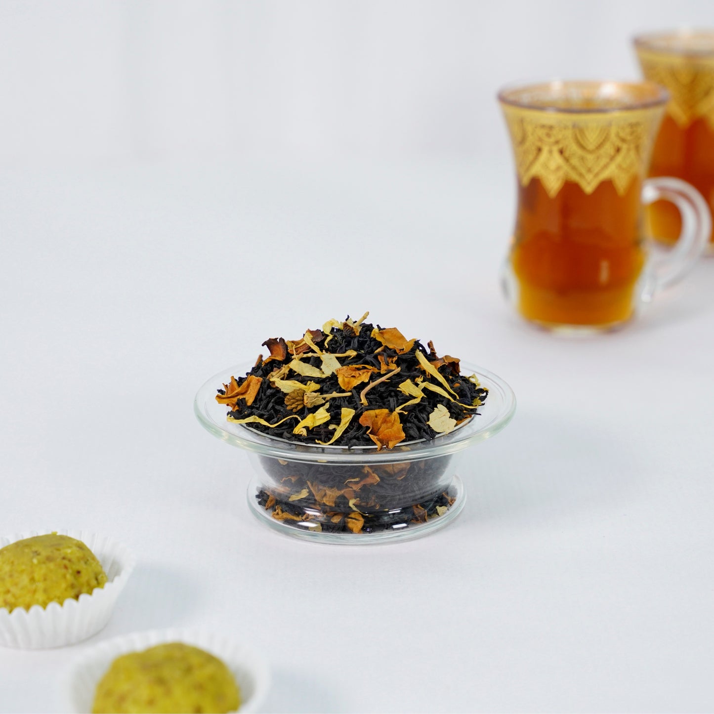 Tea leaves in a glass bowl with a cup of tea in the background on a white surface