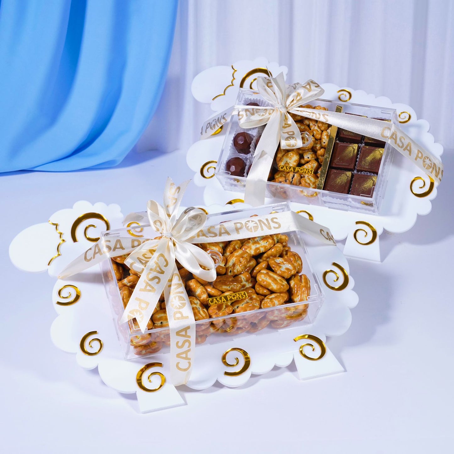 Three transparent containers filled with various snacks and chocolates, each tied with a white ribbon and 'CASA PONS' branding, on a light blue background.