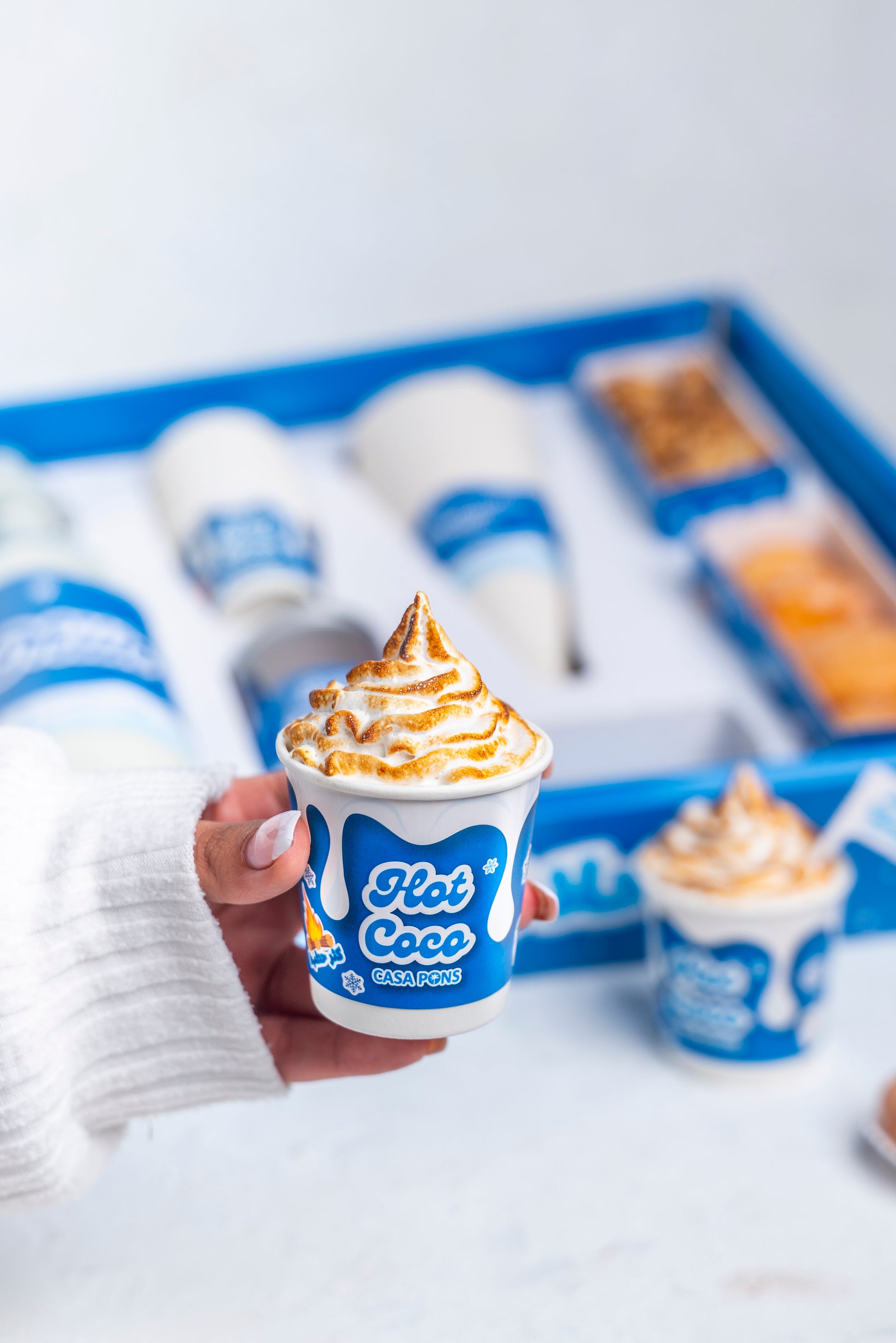 Hand holding a cup of Flat Coco dessert with a box of more desserts in the background.