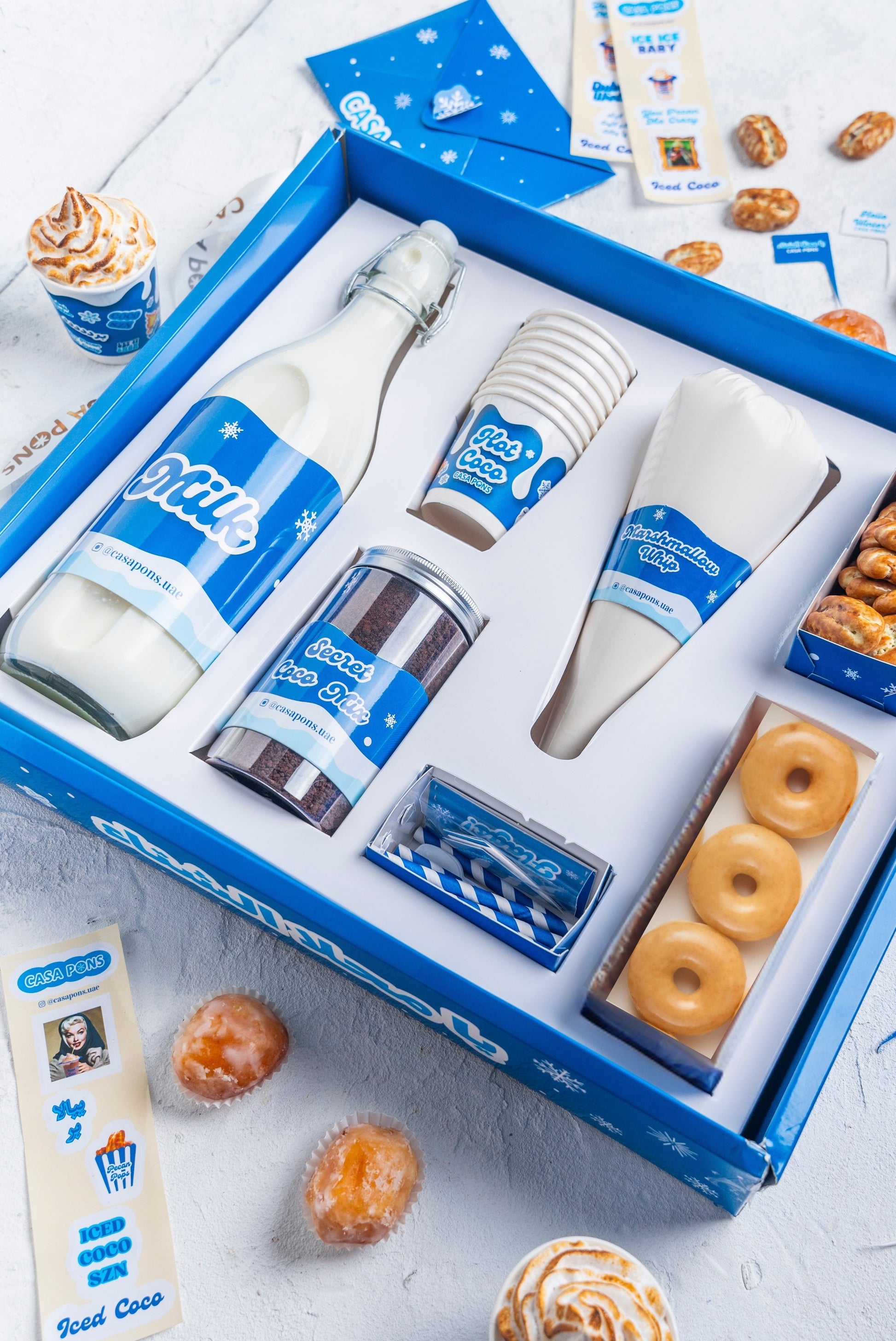 Gift set with bottles, cups, and donuts in a blue box on a light gray surface.